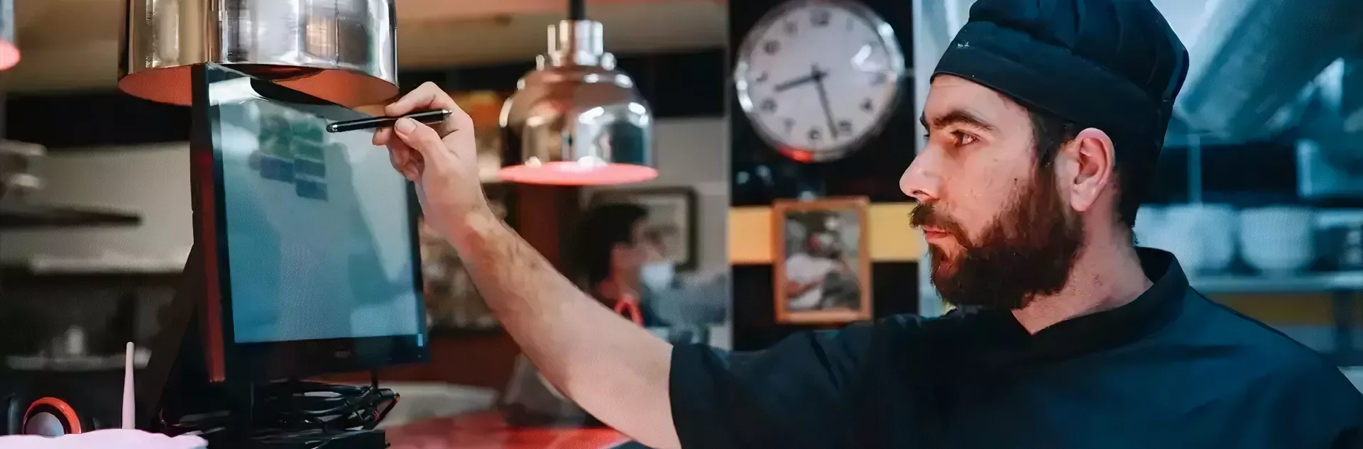 Cozinheiro com touca preta usando caneta touch para operar tela sensível ao toque em uma cozinha profissional, com pendentes metálicos e relógio ao fundo.