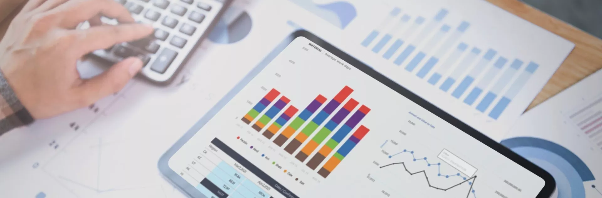 Tablet exibindo gráficos coloridos de barras e linha sobre uma mesa com planilhas e documentos de dados, enquanto uma mão opera uma calculadora ao lado.