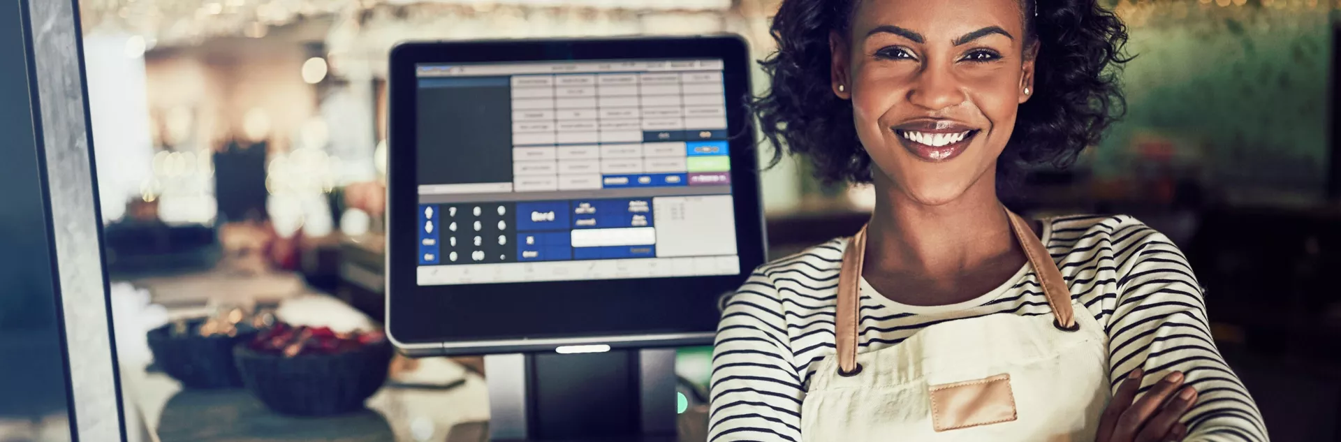 Mulher sorridente com avental claro de braços cruzados em frente a uma tela de PDV ligada, em ambiente de restaurante moderno e iluminado.