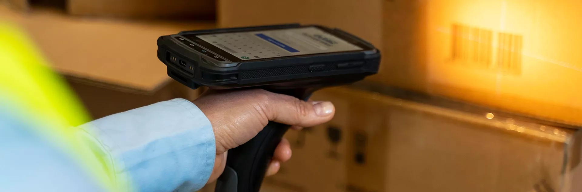 Mão segurando coletor de dados com tela acesa apontado para caixa com código de barras, em ambiente de estoque com iluminação amarela.
