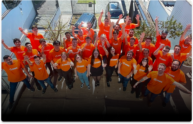 Equipe CNM reunida em área externa, usando camisetas laranja com logo da empresa, sorrindo e acenando para a câmera.