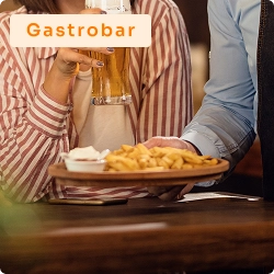 Pessoa segurando copo de chope enquanto garçom serve petiscos sobre tábua de madeira em ambiente aconchegante de gastrobar.