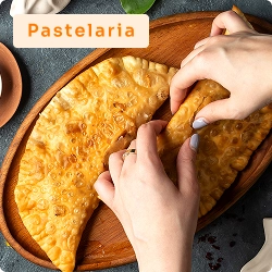 Mãos partindo pastel grande ao meio sobre prato de madeira, com acompanhamentos ao redor, representando pastelaria tradicional.