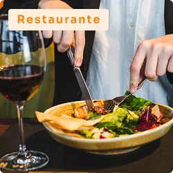 Pessoa cortando salada com garfo e faca em restaurante, ao lado de uma taça de vinho tinto, em ambiente sofisticado