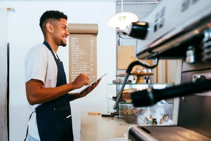 Barista sorridente anotando pedido em tablet ao lado de máquina de café, destacando a mobilidade e eficiência de um sistema para cafeterias no atendimento.